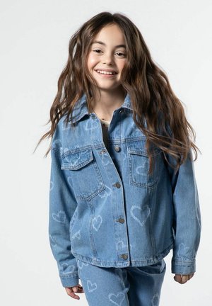 Uśmiechnięta dziewczyna z długimi brązowymi włosami, ubrana w niebieską dżinsową kurtkę i spodnie zdobione wyblakłymi wzorami serc na jednolitym tle.