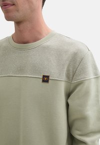 Svetlozelená mikina s diagonálnym textúrovaným prúžkovým vzorom na hornej časti, s malým oranžovým logom. Mäkkký materiál.