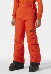 Oranžové lyžařské kalhoty volného střihu, vyrobené z voděodolného materiálu, se černým logem a nastavitelným pasem. Na spodní části jsou vidět černé lyžařské boty.