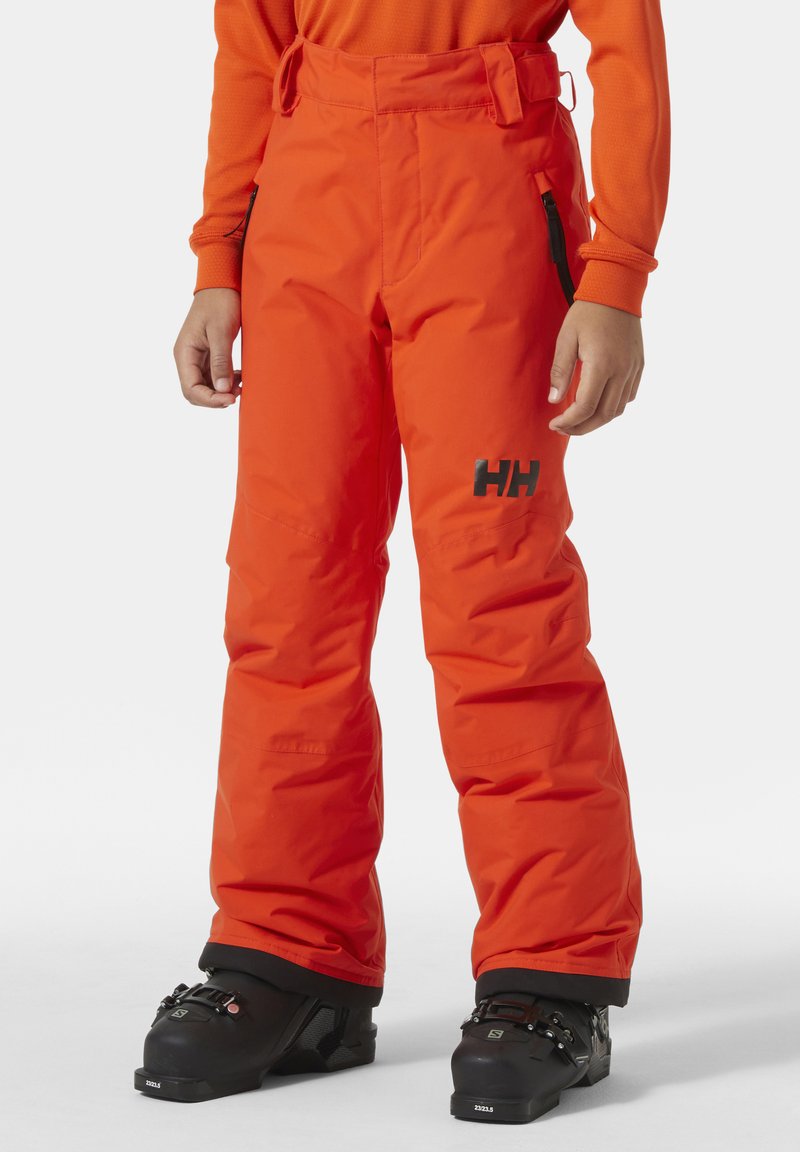 Oranžové lyžařské kalhoty volného střihu, vyrobené z voděodolného materiálu, se černým logem a nastavitelným pasem. Na spodní části jsou vidět černé lyžařské boty.