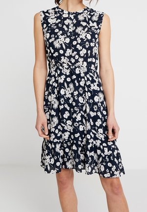 Robe bleu marine sans manches avec imprimé floral blanc, taille ajustée et ourlet à volants portée par une personne debout devant un fond uni.