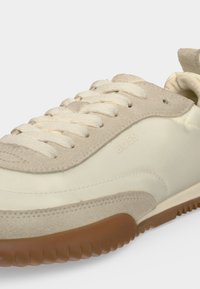 Gros plan d'une basket crème et beige avec des lacets texturés, des empiècements en daim et une semelle marron nervurée. Petit logo embossé "BOSS" visible.