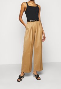 Black tank top with square neckline, high-waisted beige trousers with pleats, black belt, and open-toe black heeled sandals.