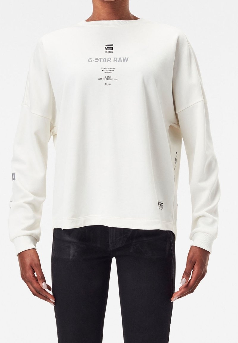 Sweat-shirt blanc à manches longues G-Star RAW porté avec un pantalon noir, avec un logo et un texte imprimés sur la poitrine et la manche.