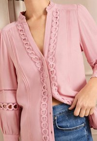 Blusa rosa con bottoni, con ritagli a forma di cuore lungo il davanti, tessuto strutturato e maniche arricciate, indossata sopra un jeans.