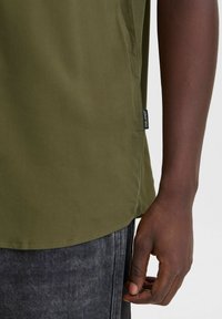 Olive green short-sleeve shirt with a straight hem, side slits, and a black tag on the lower left seam; paired with dark gray pants.