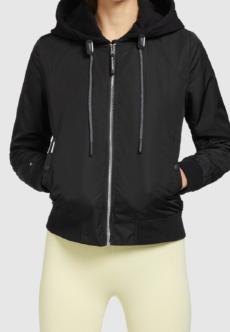 Czarna bomberka z kapturem, zapinana na zamek, z elastycznymi mankietami i sznurkami do ściągania. Posiada gładką teksturę oraz boczne kieszenie dla wygody.