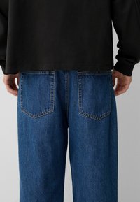 Uvoľnené modré denimové džínsy s dvoma zadnými vreckami, viditeľným šitím a mierne vyblednutou textúrou, spárované s čiernou košeľou s dlhým rukávom.