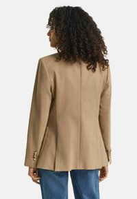Blazer in tessuto beige chiaro con silhouette dritta, due tasche laterali e dettagli in bottone dorato sui polsini. La vista posteriore mostra cuciture pulite.
