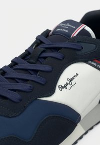 Sapatilha azul-marinho com detalhes em branco, parte superior texturizada, atacadores planos e detalhe de logótipo. Apresenta uma sola grossa e painéis de cores contrastantes.
