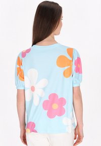 Top azzurro chiaro a maniche corte con motivi floreali sovradimensionati in rosa, arancione e bianco. Presenta un colletto rotondo e maniche arricciate.