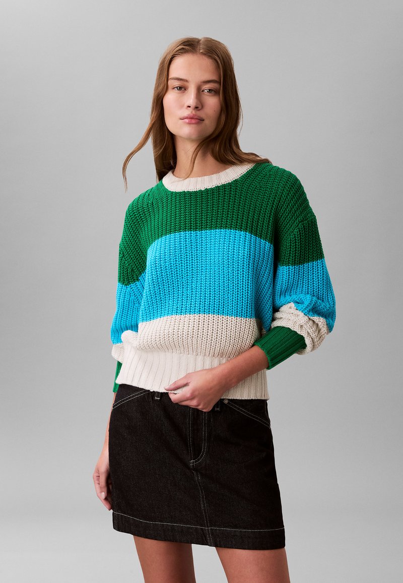 Pull en tricot avec des rayures vertes, bleues et crème, poignets et ceinture côtelés ; associé à une jupe en jean foncé avec des coutures blanches.