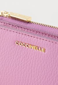 Vaaleanpunainen nahkalompakko, jossa on teksturoidu pinta, kullanvärinen vetoketju ja kohopainettu "Coccinelle"-logo. Vetoketjun kangaslaventelin värinen.