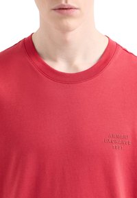 Červené tričko s krátkymi rukávmi vyrobené z mäkkej tkaniny, s jemne embosovaným logom "ARMANI EXCHANGE 1991" na ľavej strane hrude.