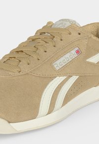 Μπεζ σουέντ αθλητικό παπούτσι Reebok με μπεζ κορδόνια, λευκές πλευρικές ρίγες και μια μικρή σημαία Union Jack κοντά στο λογότυπο της μάρκας.