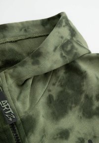 Zielona oliwkowa bluza z zamkiem błyskawicznym, ozdobiona fakturowanym wzorem tie-dye. Posiada wysoki kołnierz oraz czarną metkę z logo w pobliżu zamka. Wykonana z miękkiego materiału.