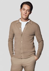 Jasnobrązowy kardigan wykonany z dzianiny, z kołnierzykiem, zapinany na guziki oraz z prążkowanymi mankietami. Noszony na białej koszuli.
