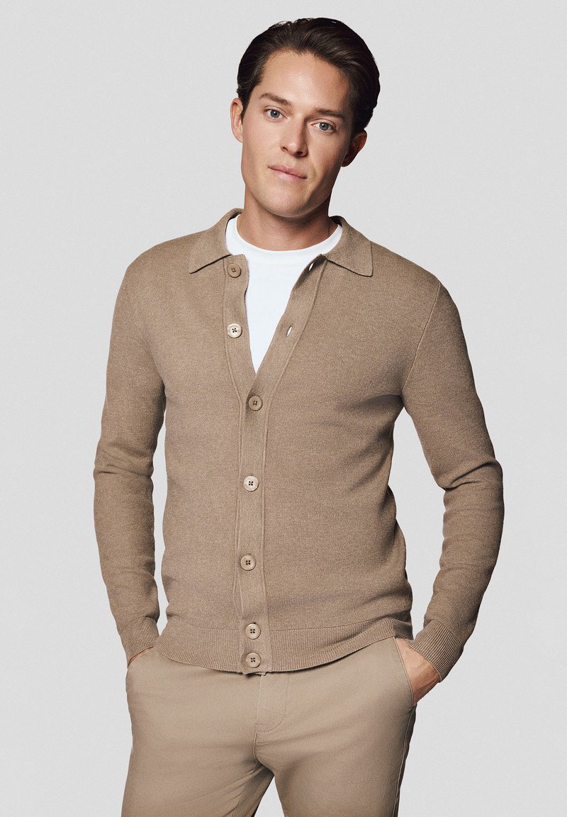Jasnobrązowy kardigan wykonany z dzianiny, z kołnierzykiem, zapinany na guziki oraz z prążkowanymi mankietami. Noszony na białej koszuli.