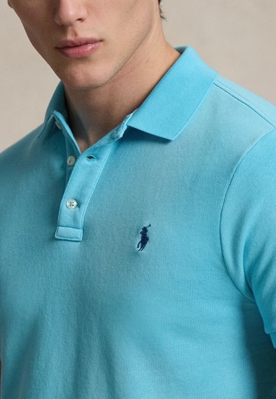 Νεαρός άντρας που φοράει ανοιχτό γαλάζιο πόλο με σκούρο μπλε κεντημένο λογότυπο αλόγου στο αριστερό στήθος.