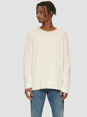 Langarm, off-white shirt met een ronde halslijn en een drieknoopsluiting; gemaakt van zacht materiaal met een relaxte pasvorm. Gepaard met blauwe jeans.