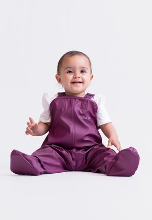 Lila wasserdichte Baby-Latzhose mit verstellbaren Trägern und einem Frontlogo. Getragen über einem weißen kurzärmeligen Shirt, sitzend auf einem hellen Hintergrund.