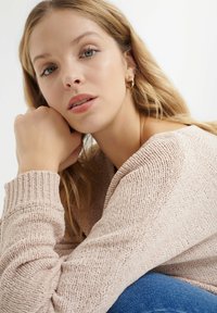 Light pink knitted sweater with ribbed cuffs and a V-neck design. Model has beige skin, light brown hair, and is seated, resting chin on hand.