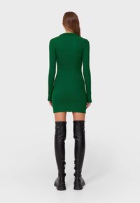 Mini-robe verte à manches longues avec une coupe ajustée. Bottes noires brillantes montant jusqu'en haut des cuisses. Texture de tissu lisse, col simple et style minimaliste.