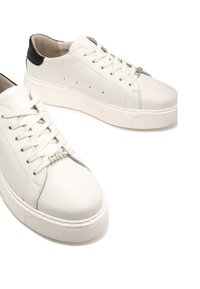 Białe skórzane trampki mają grubą podeszwę, czarny akcent na pięcie, teksturowane sznurowadła i subtelne perforacje po bokach. Widoczny detal w postaci srebrnego logo.