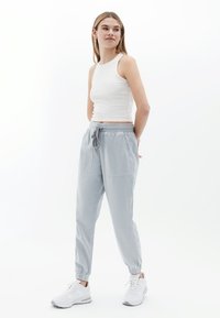 Hellegraue, elastische Bundhose mit zwei vorderen Taschen, mit Bündchen an den Knöcheln, kombiniert mit einem weißen, taillierten Tanktop und weißen Sportschuhen.