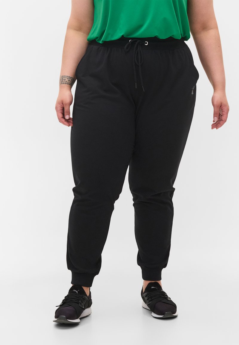 Pantaloni sportivi neri con vita regolabile tramite cordino, con polsini elasticizzati e tasche laterali. Tessuto morbido con una texture liscia. Abbinati a sneakers nere.