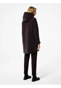 Cappotto impermeabile marrone scuro con cappuccio, caratterizzato da una linea dritta, maniche sartoriali e dettagli neri a contrasto su polsini e pantaloni.