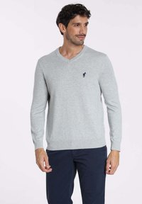 Pull en V gris clair en maille douce avec des poignets et un ourlet côtelés. Présente un petit logo de cheval bleu marine sur la poitrine gauche.