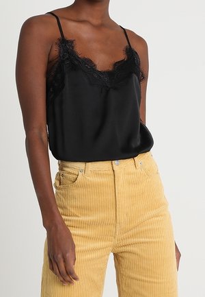 Femme portant un caraco en satin noir bordé de dentelle, rentré dans un pantalon en velours côtelé jaune taille haute, debout devant un fond uni.