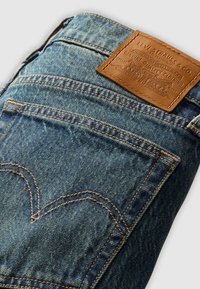 Blå denimjeans med en brun läderlapp som har präglad märkning, ljus sömnad och en hjärtformad bakfickedesign.