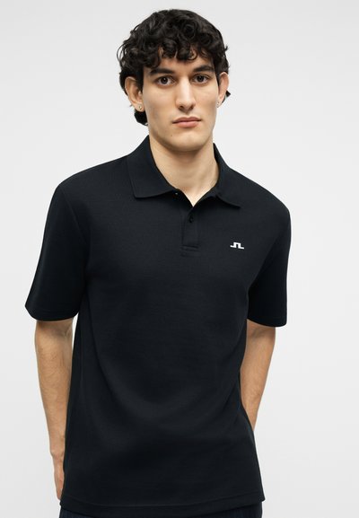 Νεαρός άνδρας με καστανά σγουρά μαλλιά, φορώντας μαύρο πόλο με κοντά μανίκια και ένα μικρό λευκό λογότυπο στο στήθος, στέκεται μπροστά σε απλό φόντο.