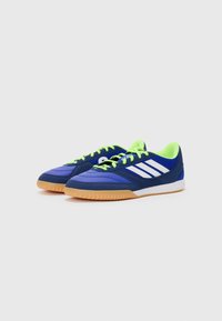 Zapatillas deportivas azules con cordones verdes, parte superior de ante y malla, rayas blancas y suela de goma. Diseño de perfil bajo para deportes en interiores.