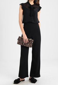 Blusa nera con maniche a ruches e dettagli a fiocco, abbinata a pantaloni neri a vita alta. Tiene una pochette con stampa leopardata e indossa mule nere.