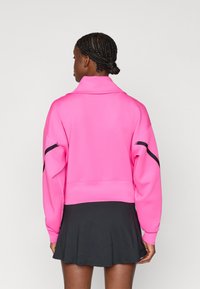 Ροζ κοντό φούτερ με ψηλό γιακά, που διαθέτει μαύρες διακριτικές ρίγες στα μανίκια, σε συνδυασμό με μια μαύρη φούστα με φάσες.