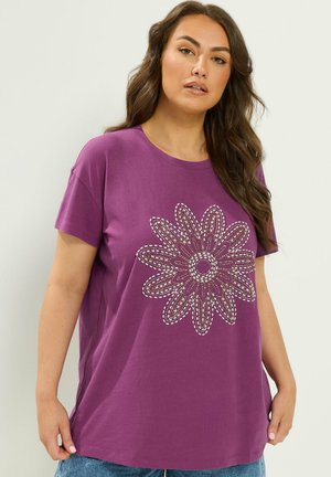 Kvinde iført en lilla kortærmet skjorte med et stort blomsterperle-design foran og blå jeans mod en ensfarvet baggrund.
