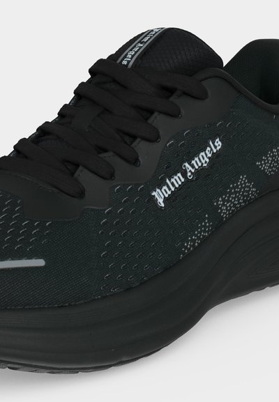 Κοντινή λήψη μαύρου sneaker Palm Angels με υφασμάτινη υφή, επενδυμένη γλώσσα, μαύρα κορδόνια και χοντρή σόλα.