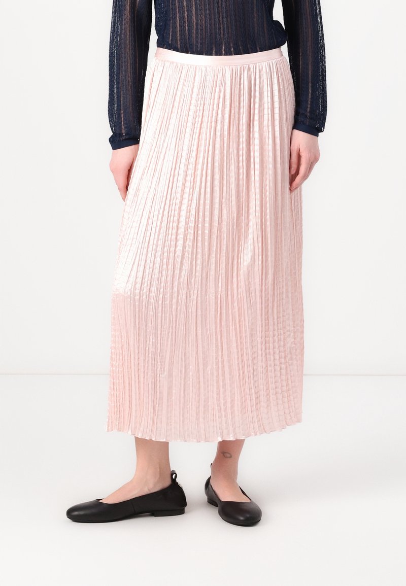 Femme portant une jupe midi plissée rose clair, un haut transparent bleu foncé à manches longues, et des chaussures plates noires, debout sur un sol blanc.