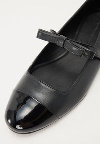 Sabot din piele neagră, cu vârful rotunjit, având un vârf lucios negru, un superior texturat și un accent sub formă de funda pe curea.