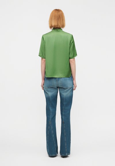 Πράσινο σατέν πουκάμισο με κοντά μανίκια, σε μήκος cropped και με ίσια κοπή. Φορεμένο με μπλε τζιν καμπάνα που διαθέτει ραφές ως διακοσμητικά στοιχεία.
