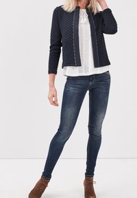 Mujer vistiendo una chaqueta de cuadros azul marino, blusa blanca, jeans skinny azul oscuro y botas de ante marrón, de pie contra un fondo liso.