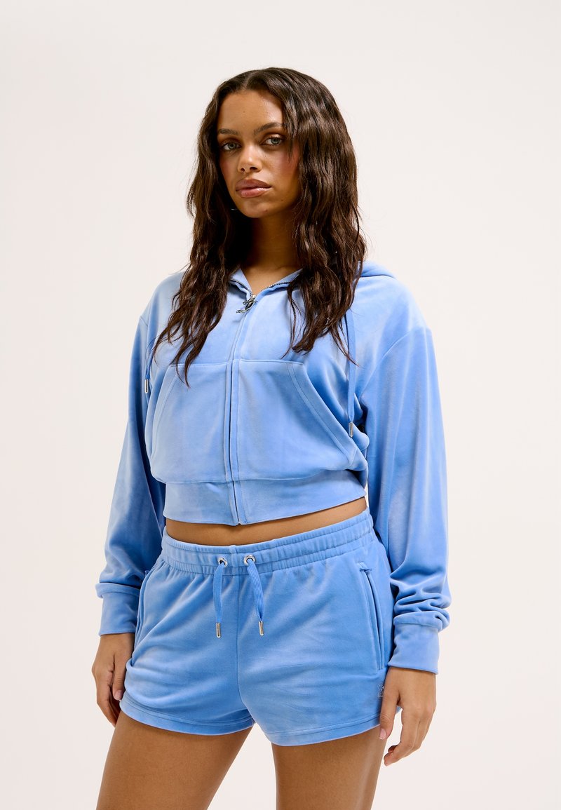 Felpa zip-up in velluto di colore azzurro chiaro con design cropped e pantaloni corti abbinati con vita elasticizzata, dotati di tasche laterali e dettagli con cordoncino.
