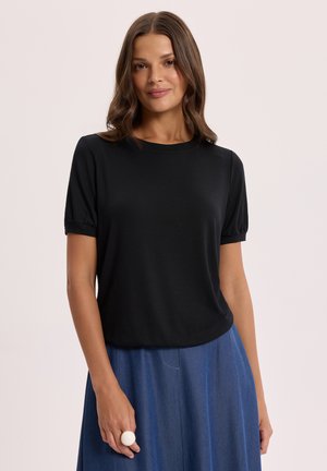 Kobieta o brązowych włosach do ramion, ubrana w czarną bluzkę z krótkim rękawem i niebieską spódnicę, stojąca na tle jasnego, jednolitego tła.