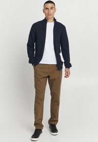 Marinblå stickad zip-jacka med texturerade axlar, tillsammans med en vit t-shirt och bruna byxor. Svarta sneakers kompletterar outfiten.