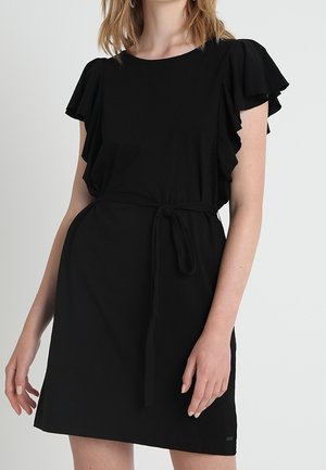 Femme portant une robe noire à manches courtes avec des manches volantées et une ceinture nouée à la taille, debout devant un fond clair uni.