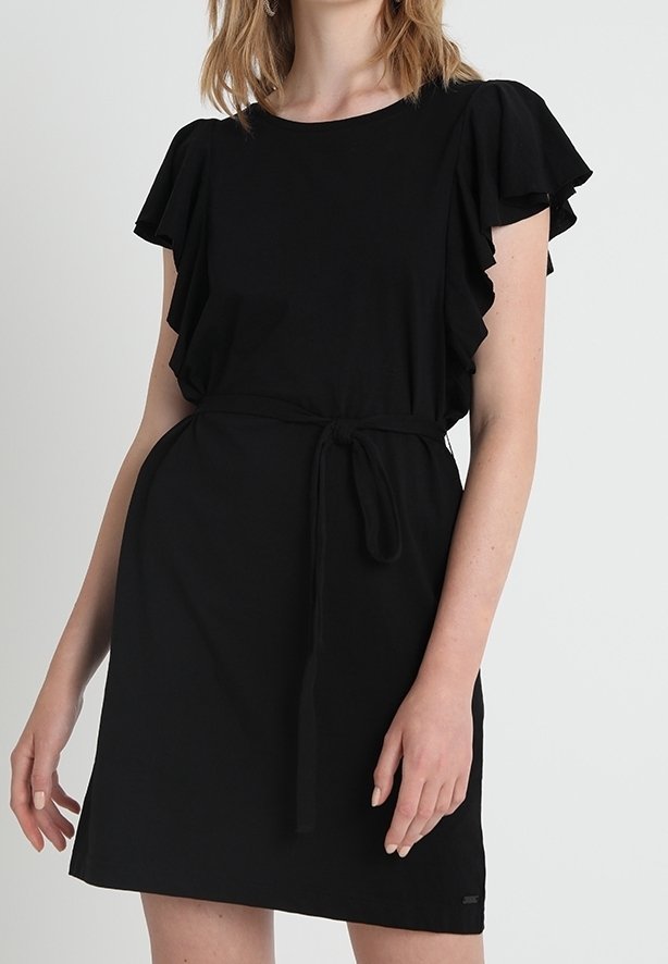 Femme portant une robe noire à manches courtes avec des manches volantées et une ceinture nouée à la taille, debout devant un fond clair uni.