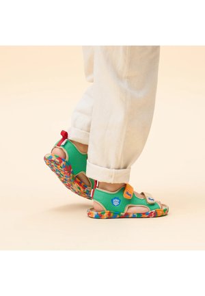 Sandalias verdes con correas ajustables, con una suela de patrón multicolor y un logo de marca. Pie bronceado visible, usadas con pantalones blancos.
